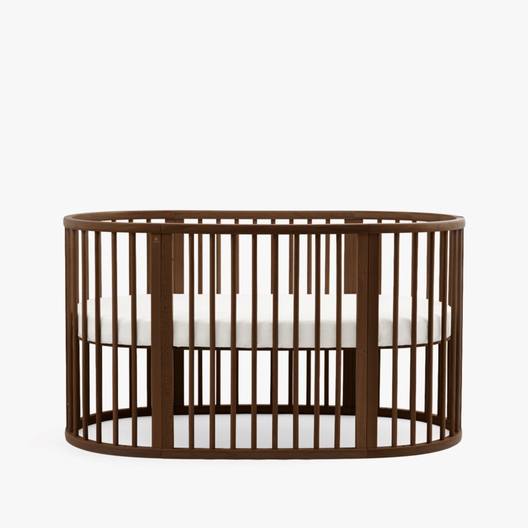 En brun oval babyseng med vertikale spiler og en hvit madrass.