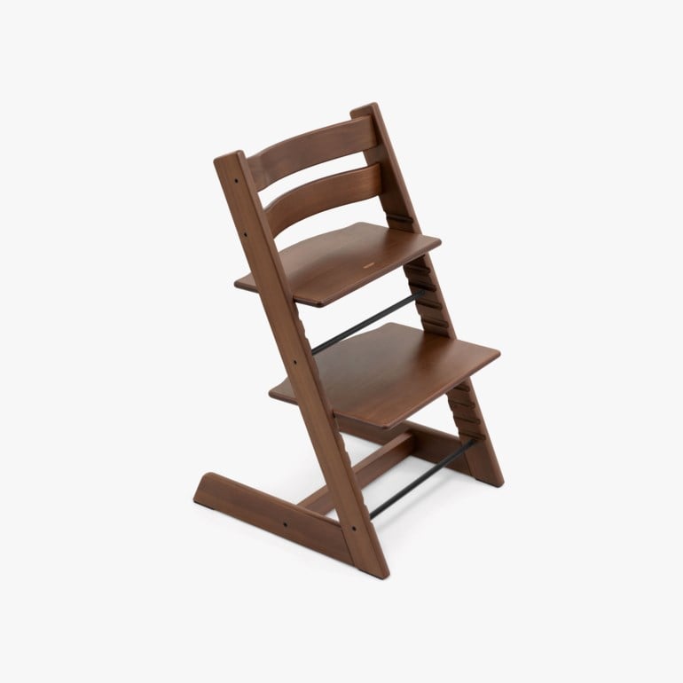 Brun barnestol i tre med justerbart sete og fotstøtte, med svarte metallstenger, sett fra skrå vinkel.