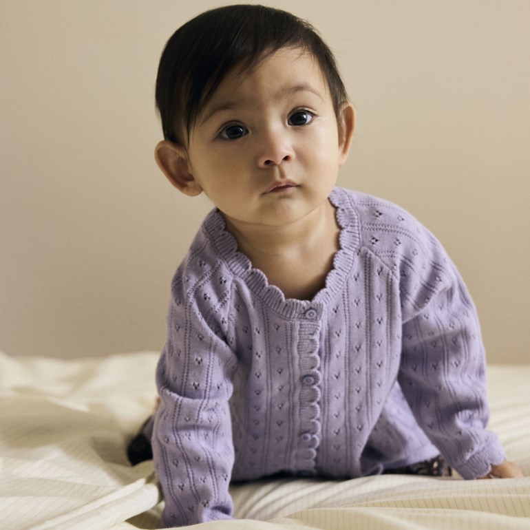 Frontvisning av baby i lys lilla strikket cardigan med bølgete kanter og perforert mønster.