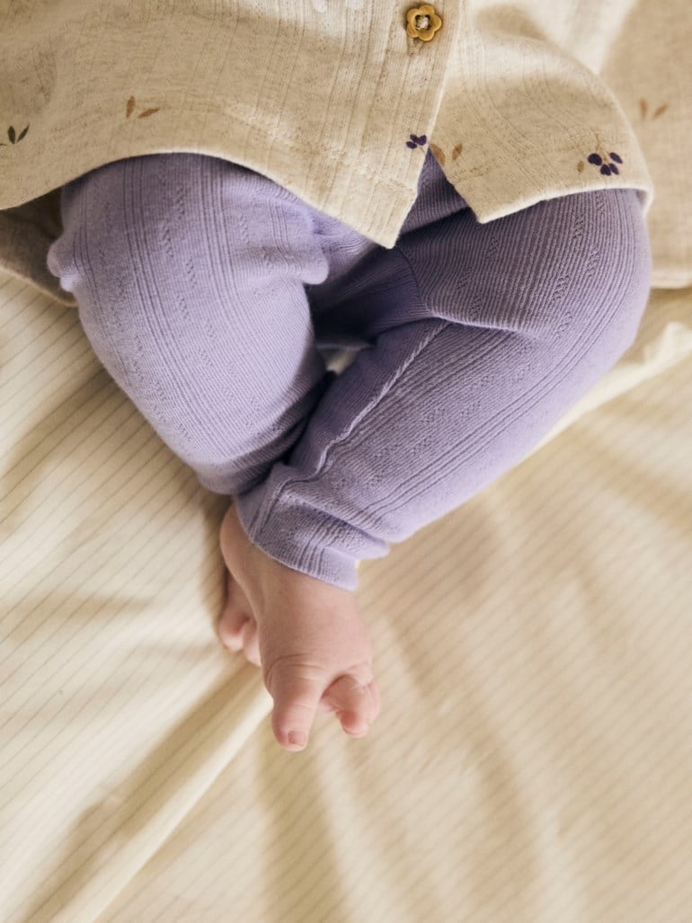 Nærbilde av baby i lys lilla mønstrede leggings og en beige blomstret cardigan med en blomsterknapp.