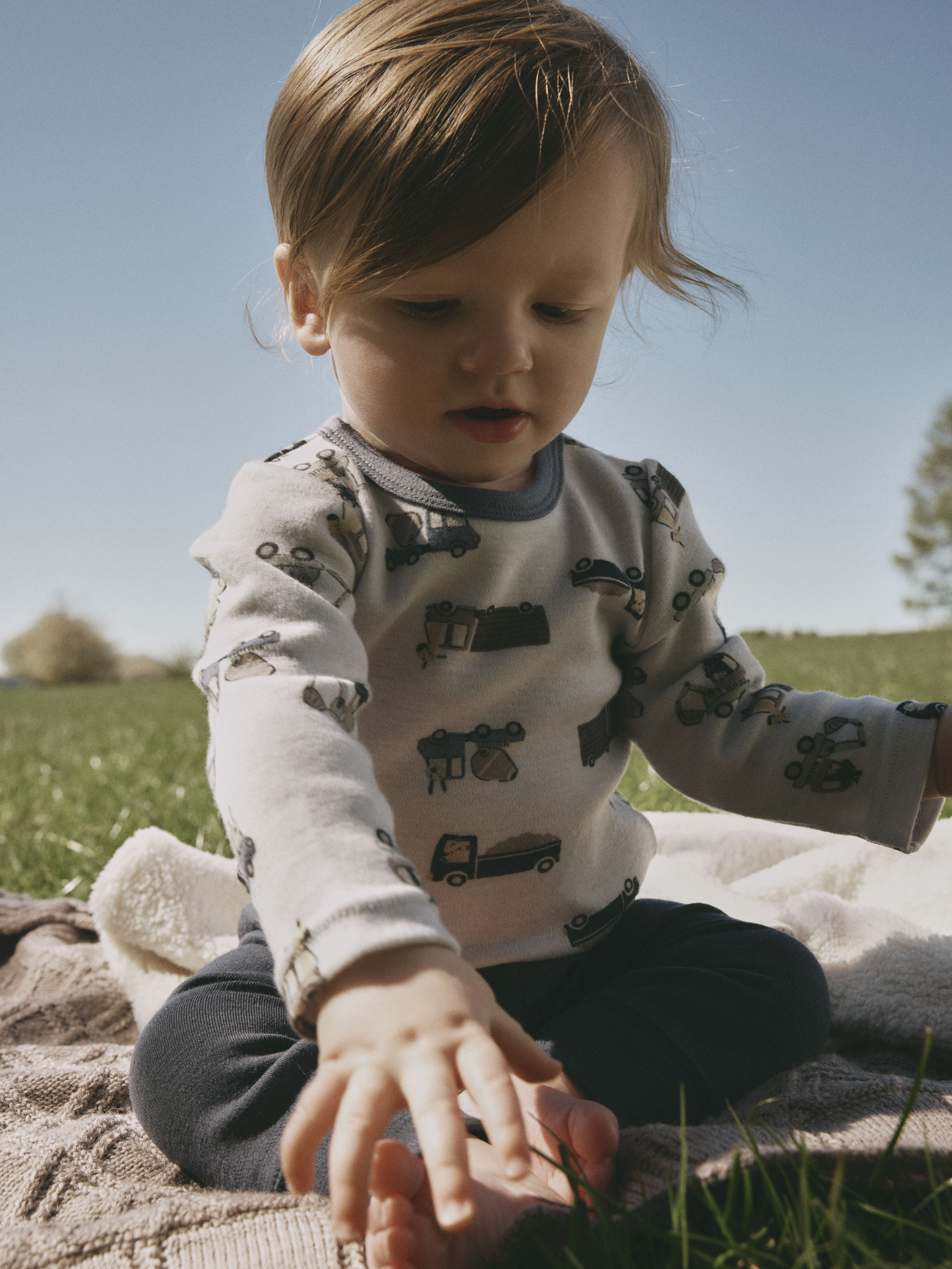 Baby har på seg lys grå langermet topp med kjøretøymønster og mørk grå bukse, sitter på et teppe.