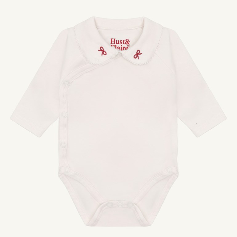 Hvit langermet babybody med bølget krave, røde broderte sløyfer og trykknappfester.