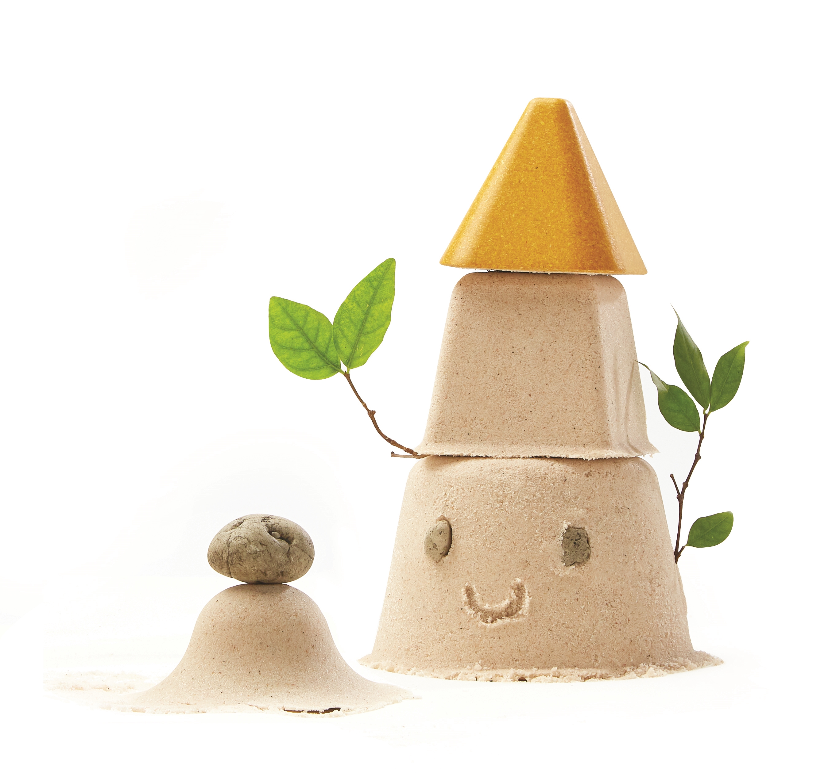 Staplet sandslott med gul topp, grønne blader og smilende ansikt, ved en liten sandhaug med stein.