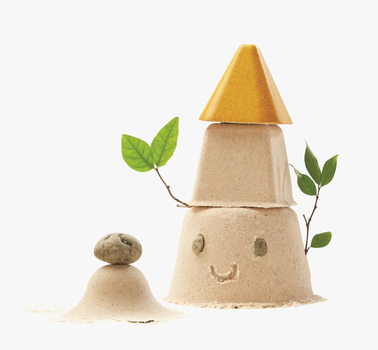 Staplet sandslott med gul topp, grønne blader og smilende ansikt, ved en liten sandhaug med stein.