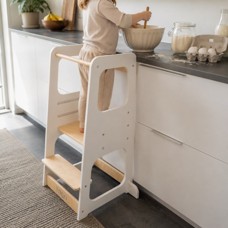 Hvit læringstårn i tre med naturfargede trinn, brukt av et barn i beige ribbet topp og bukser.