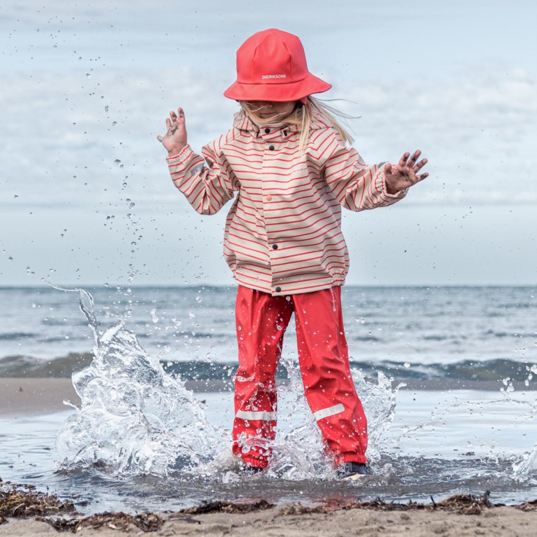 Et barn i røde regnbukser med refleksstriper, stripete regnjakke og rød regnhatt plasker i vann.