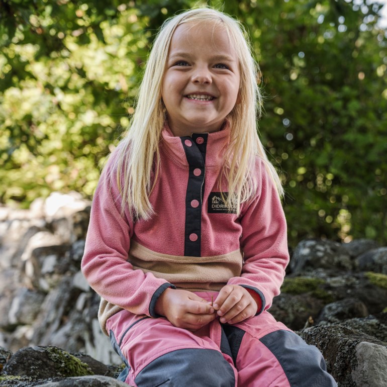 Barn iført en rosa og beige fleecetrøye med trykknapper og rosa/grå utebukse, holder en kvist.