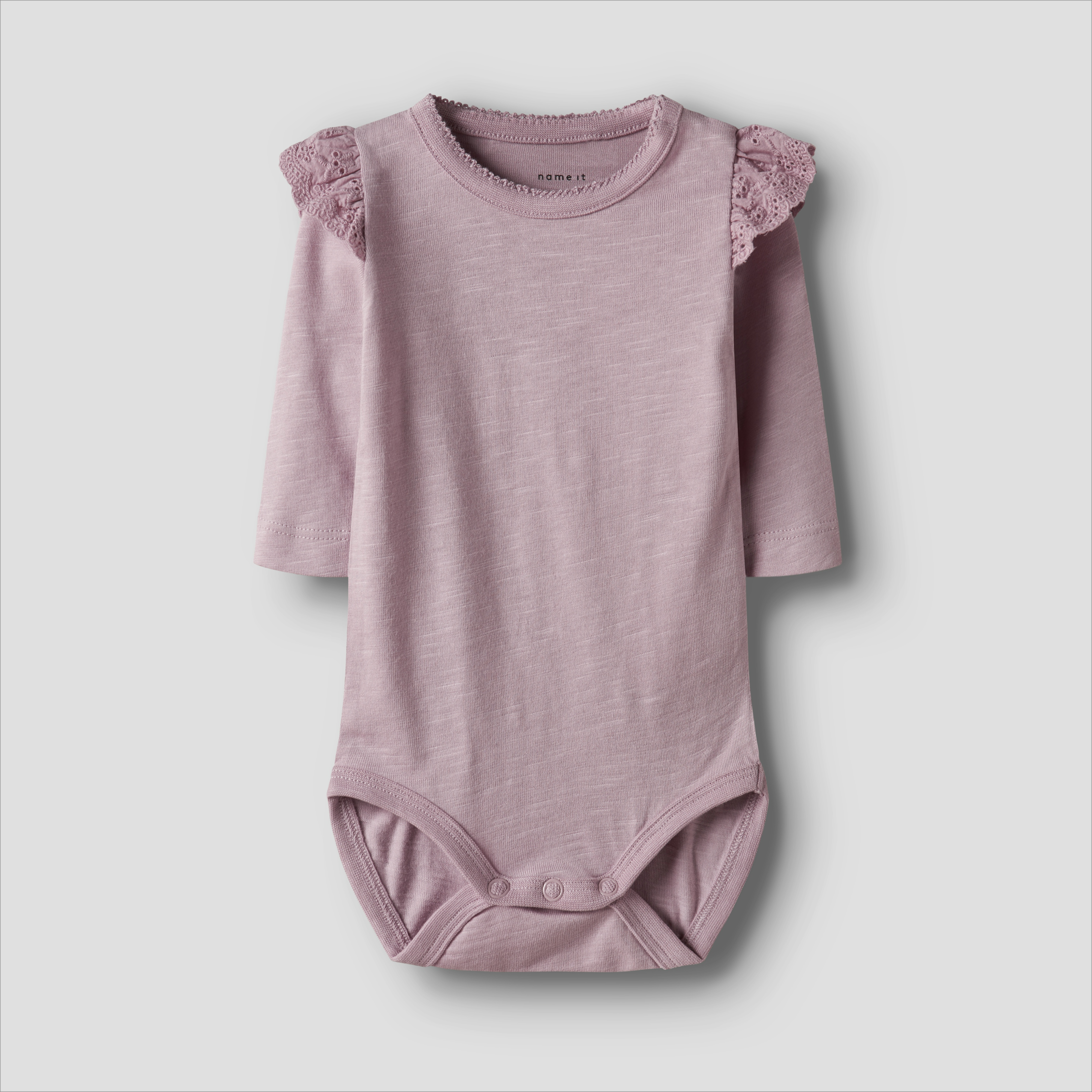 En baby lys lilla langermet body med blondevolanger på skuldrene og trykknapper, forfra.