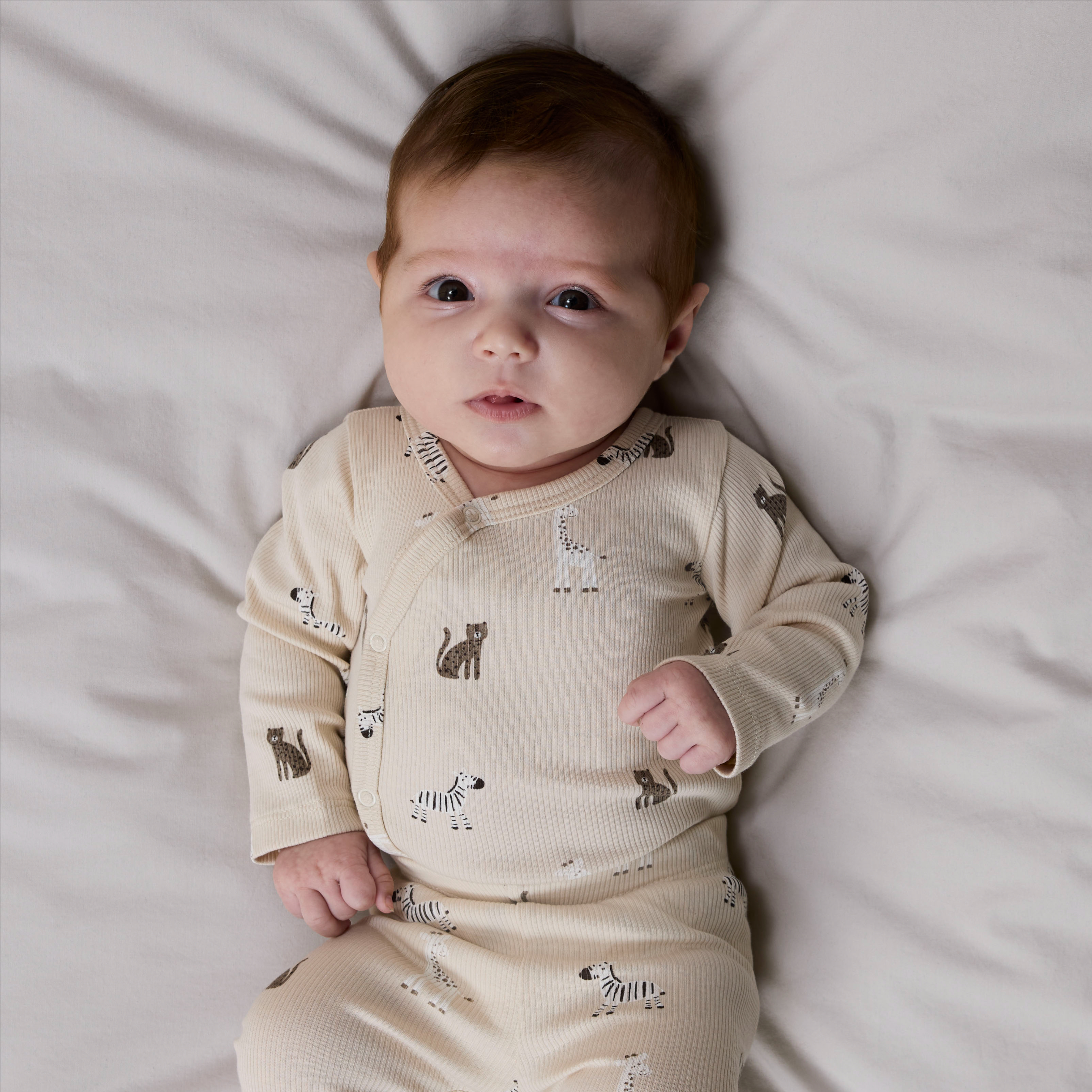 Baby har på seg en lys beige ribbet heldress med dyremønster og omslagsknapping samt lysegrå sokker.