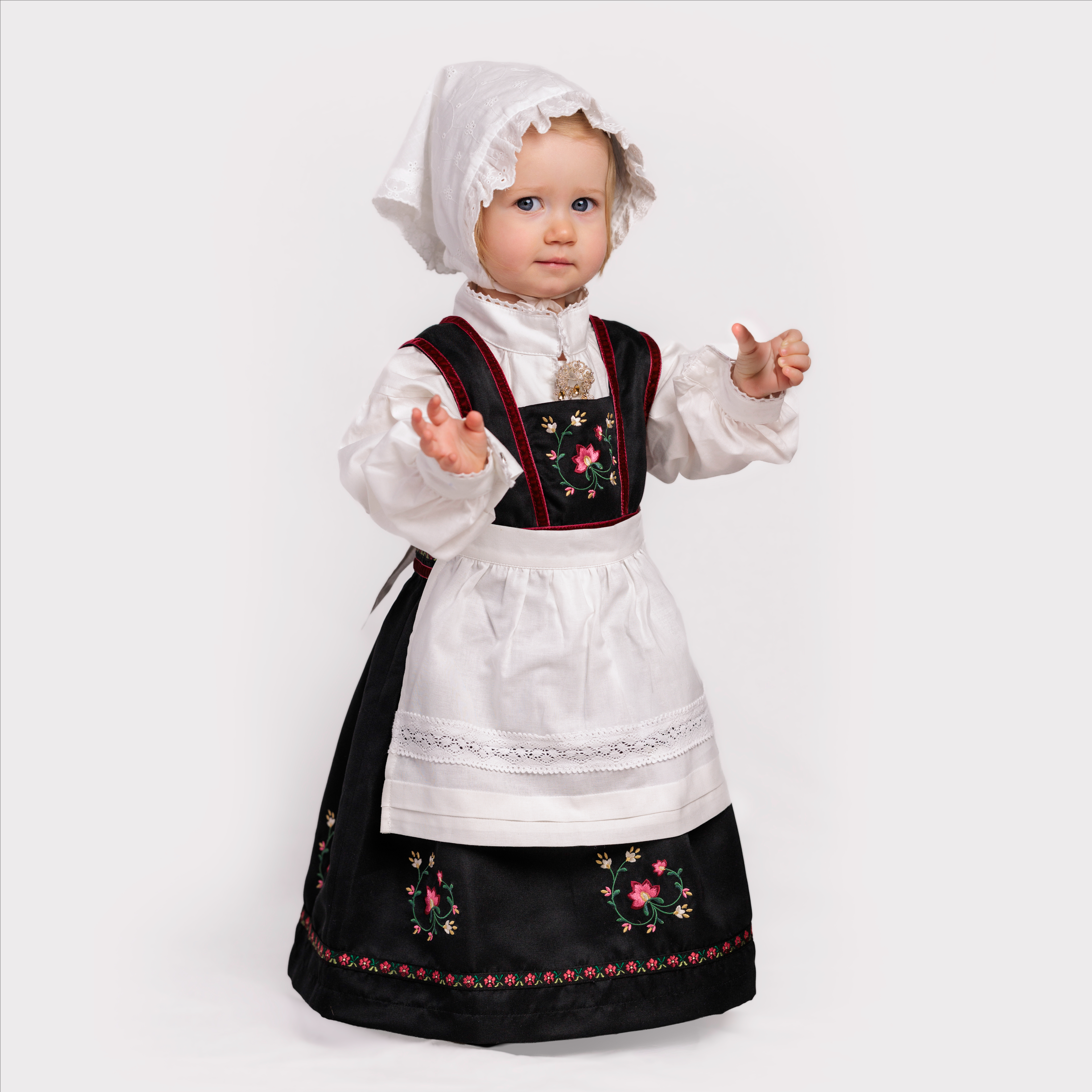 Baby bærer en tradisjonell svart bunad med blomsterbroderier, hvit skjorte, forkle og lue.