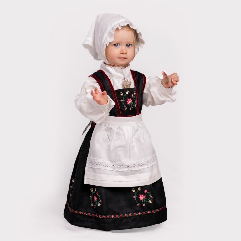Baby bærer en tradisjonell svart bunad med blomsterbroderier, hvit skjorte, forkle og lue.