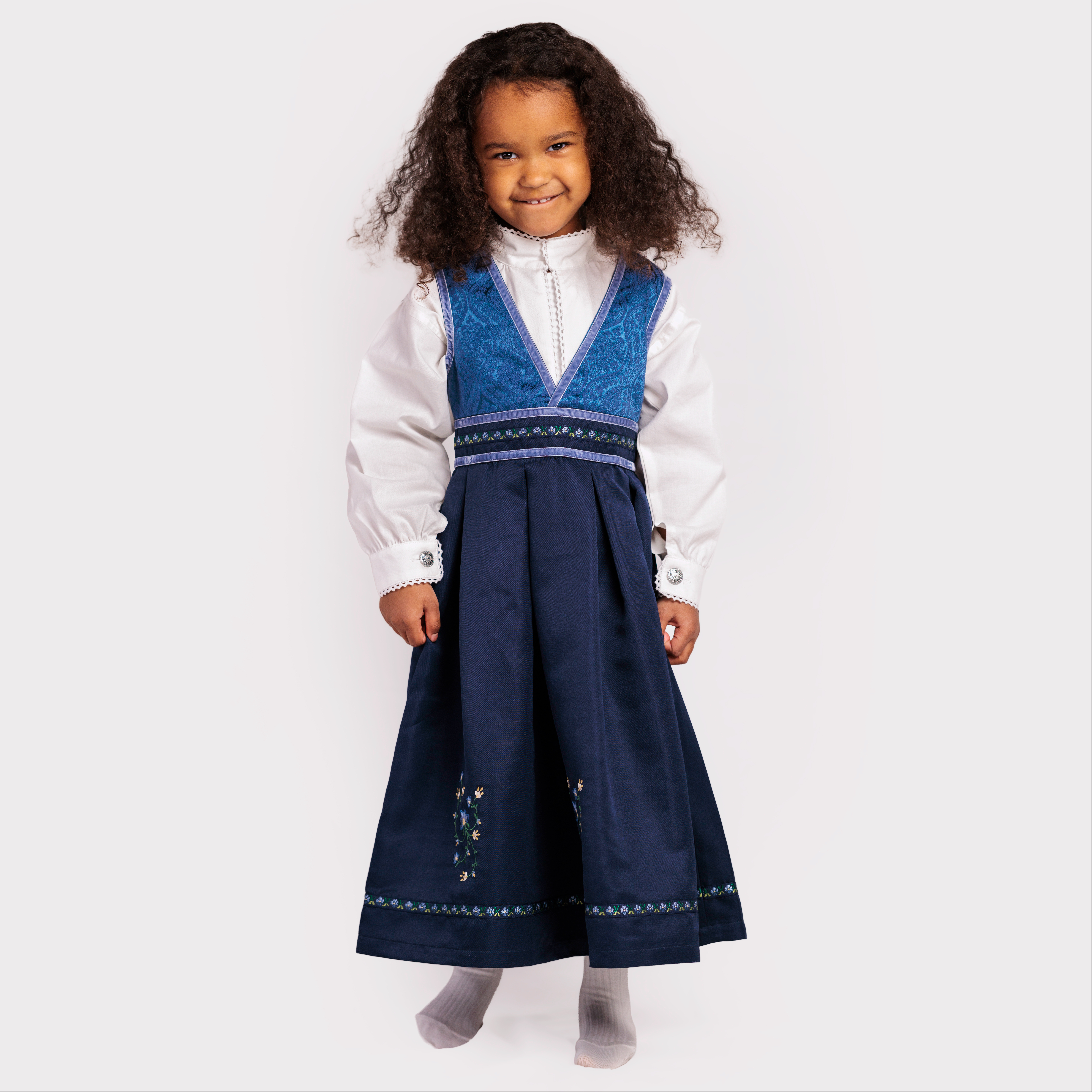Barn i mørkeblå bunad med plissert skjørt og blomsterbroderi, blå vest, hvit skjorte og grå sokker.