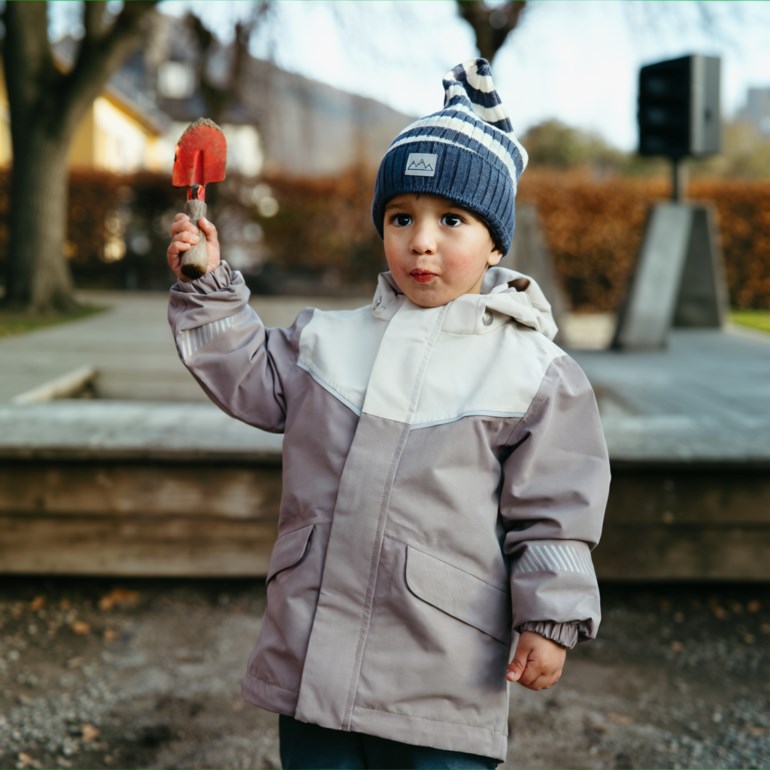 Barn har på seg en tofarget grå og beige hettejakke og en blå- og hvitstripet lue, holder en rød spade.