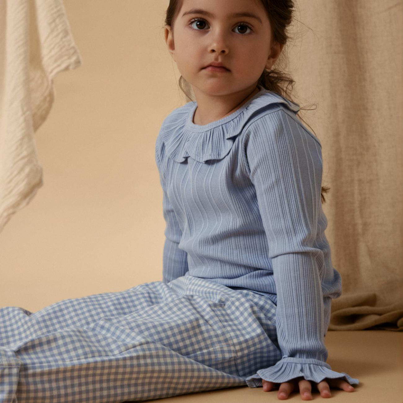 Barn i lys blå ribbestrikket langermet topp med rysjekrage og mansjetter, og blårutete bukser.