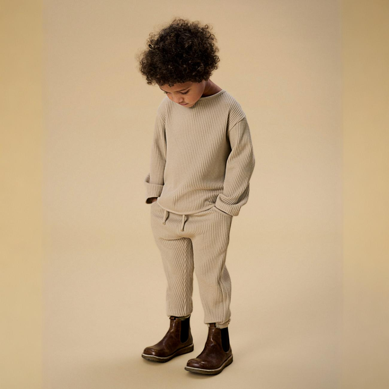 Barn i beige vaffelmønstret langermet genser, matchende bukser med elastisk midje og brune støvler.