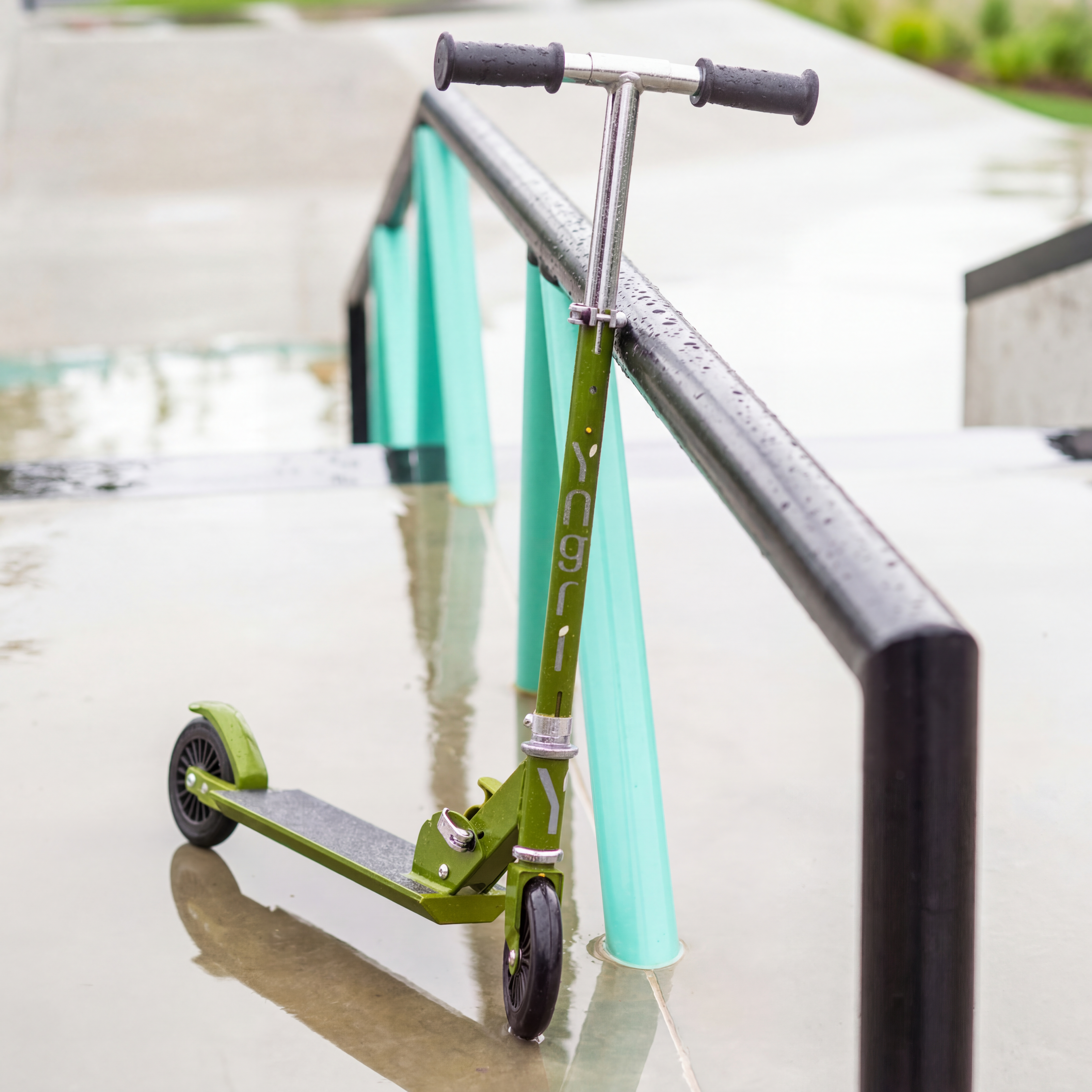 Grønn sparkesykkel med svarte hjul lener seg mot et metallrekkverk på en våt overflate.
