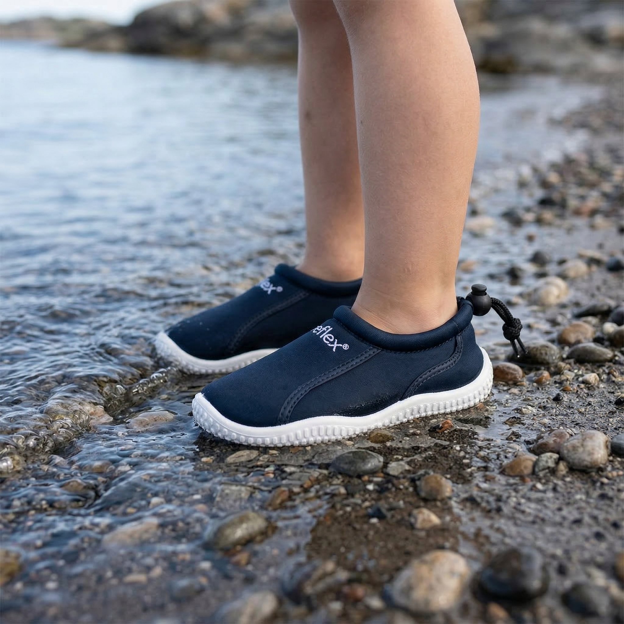 Et barn har på seg mørkeblå vannsko med hvite såler og en svart snøring, stående i grunt vann.