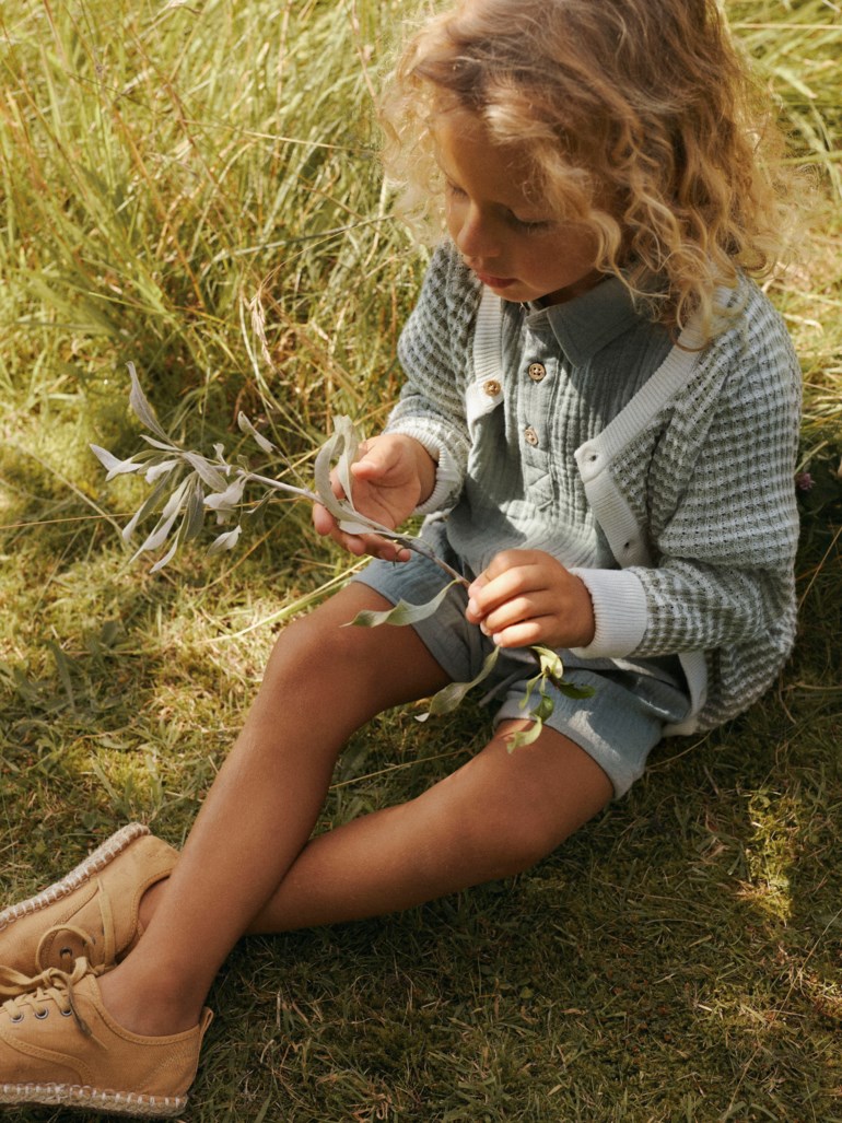 Barn bærer en grønn-hvit strikket cardigan, lysegrønn skjorte, lyseblå shorts og beige snøresko.