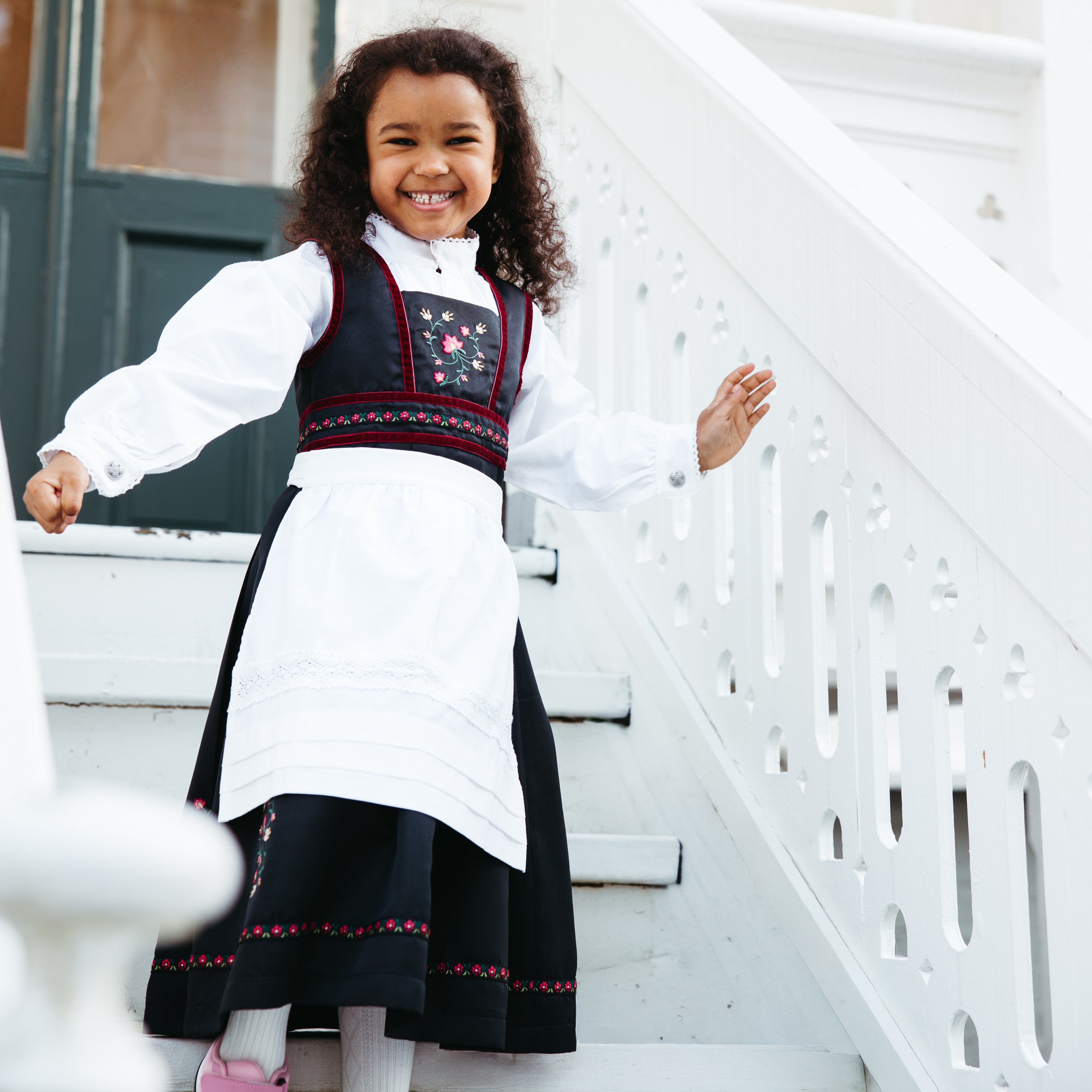 Barn i bunad med svart brodert vest, hvit bluse, svart skjørt med blomsterkant, hvitt forkle og rosa sko.