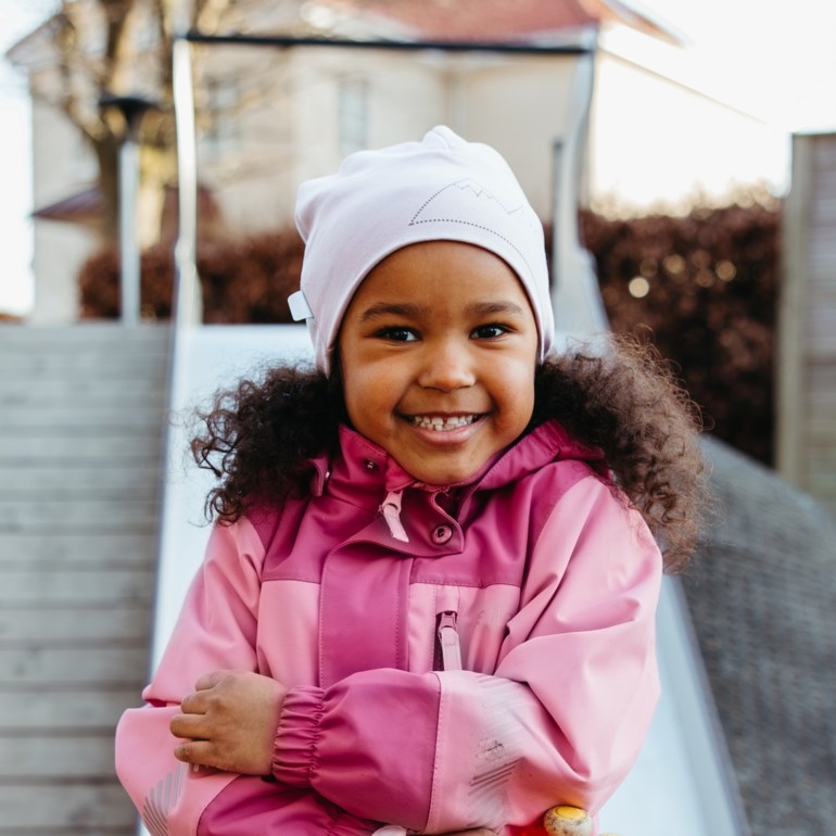 Frontbilde av et barn i en rosa og fuksia fargeblokk jakke og en lys rosa lue med fjellgrafikk.