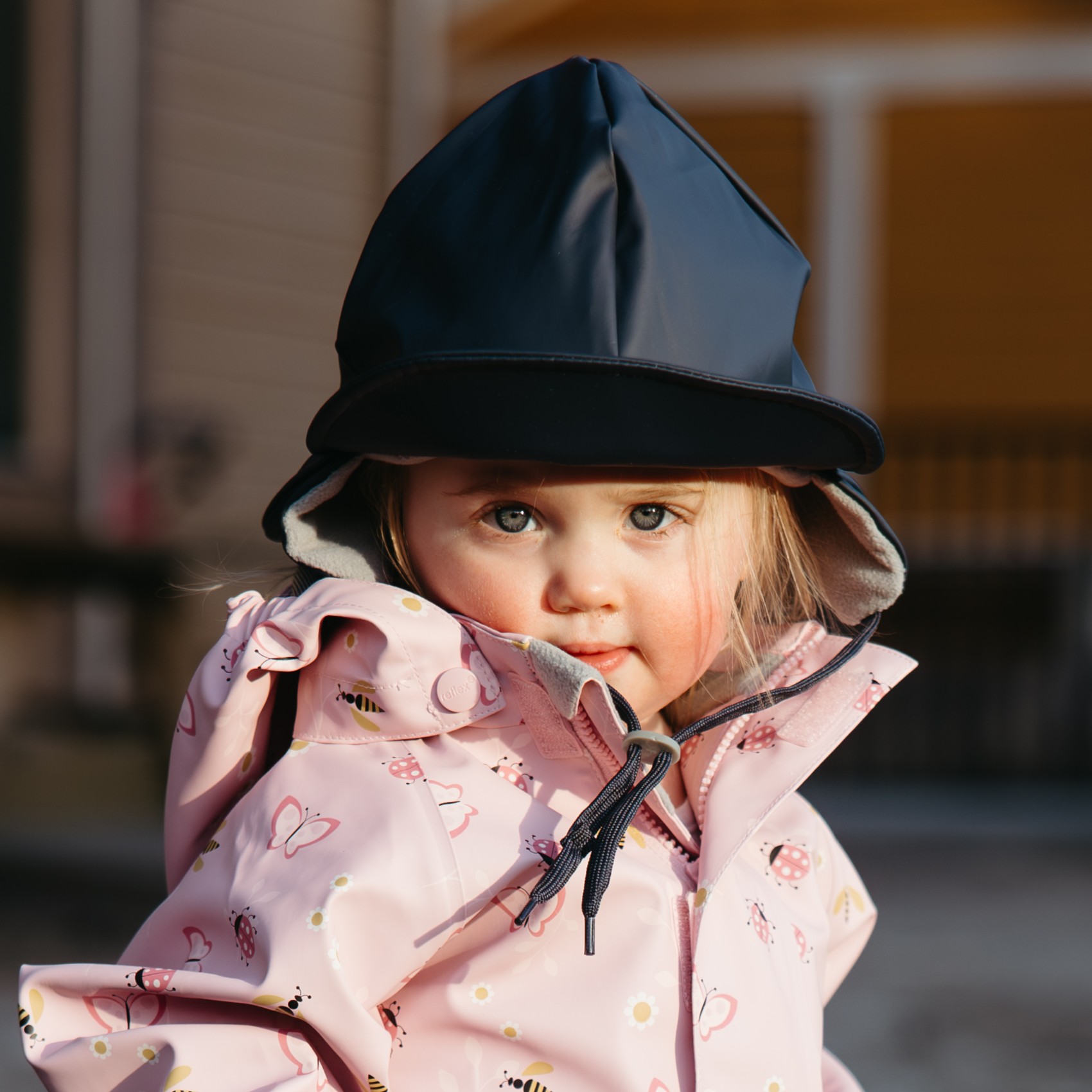 Barnet har på seg en rosa mønstret regnjakke med glidelås og snøring, samt mørkeblå regnhatt.