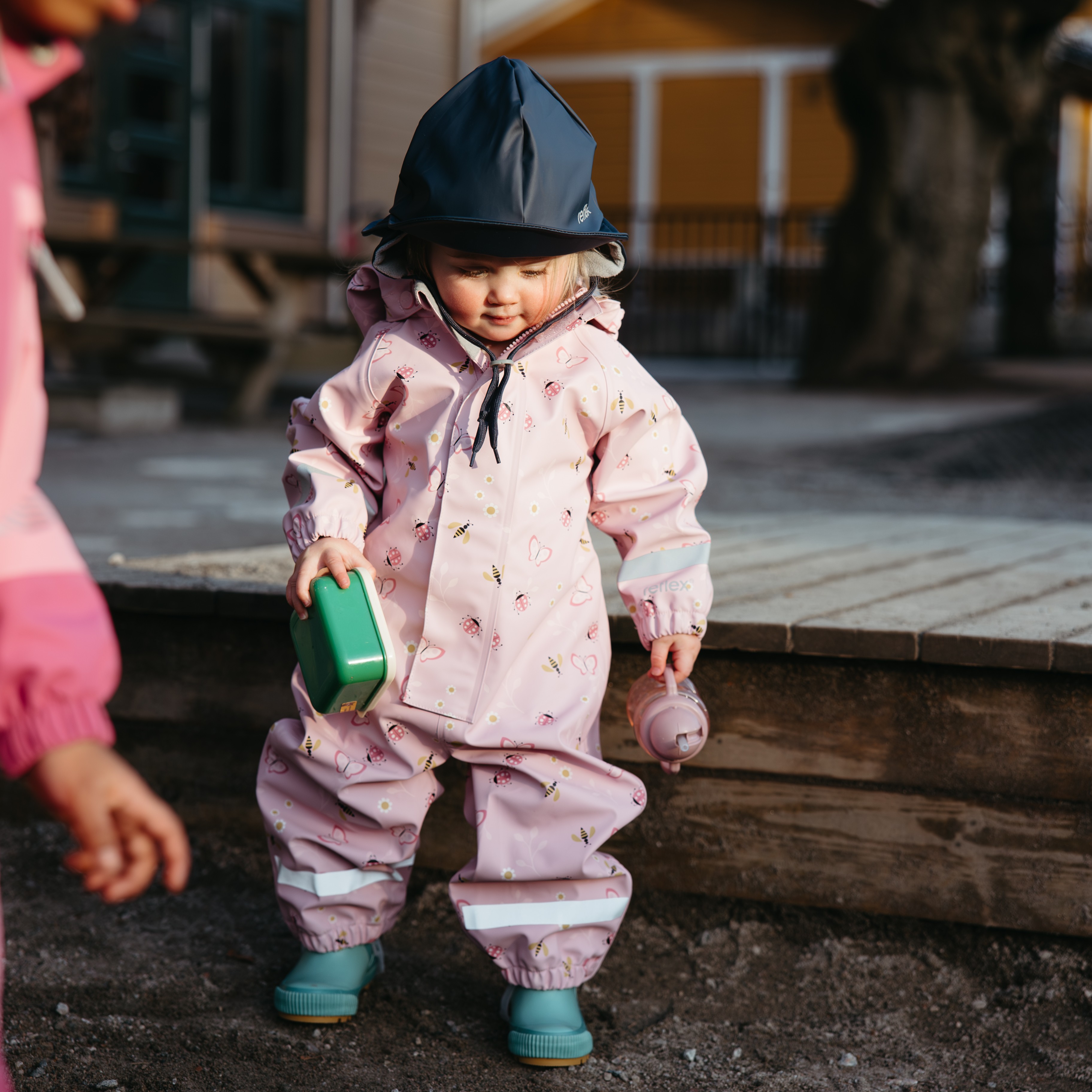 Barn har på rosa mønstret regndress, mørkeblå hatt, lyseblå støvler, holder grønn beholder og rosa kopp.