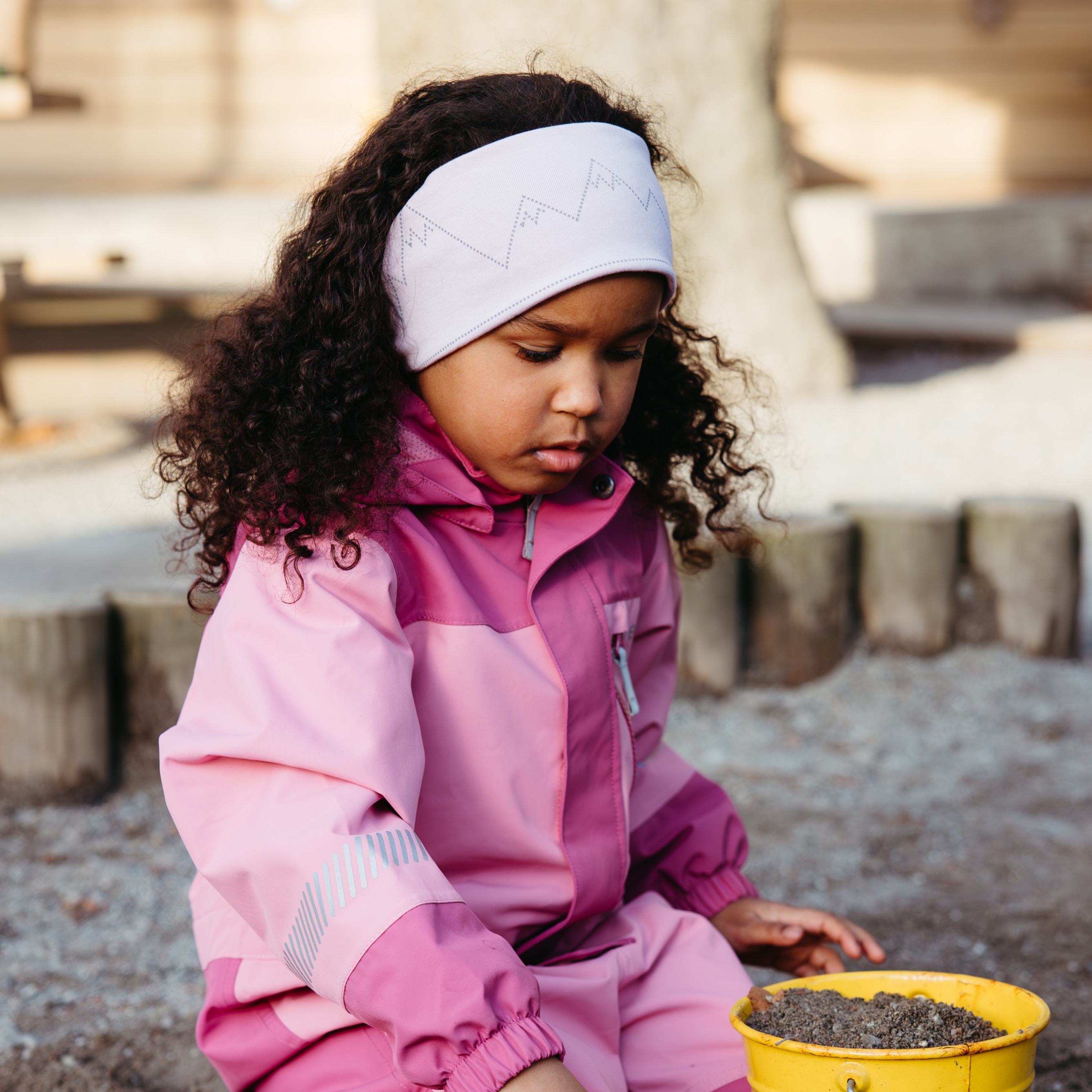 Barn i rosa vanntett dress med refleksstriper, hvitt fjellpannebånd, ser på jord i gul bøtte.