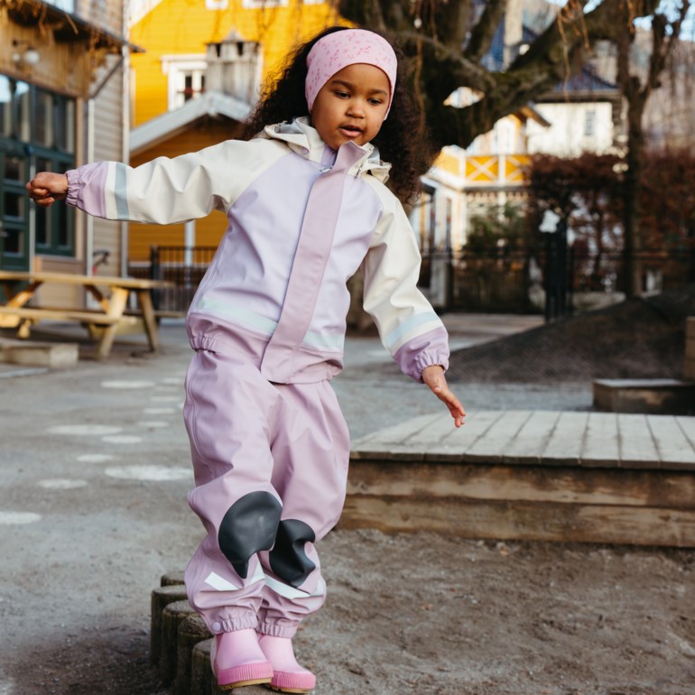 Barn i lilla og beige regndress med svarte knepolstringer, rosa pannebånd og rosa gummistøvler.