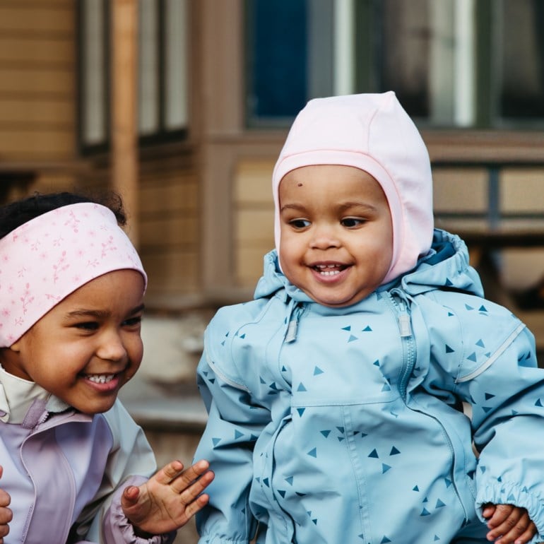 Barn i lyseblå trekantet dress og rosa balaklava; annet barn i rosa blomstret pannebånd og lilla jakke.
